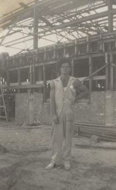 Worker wearing slouch hat and overalls in front of Mill construction