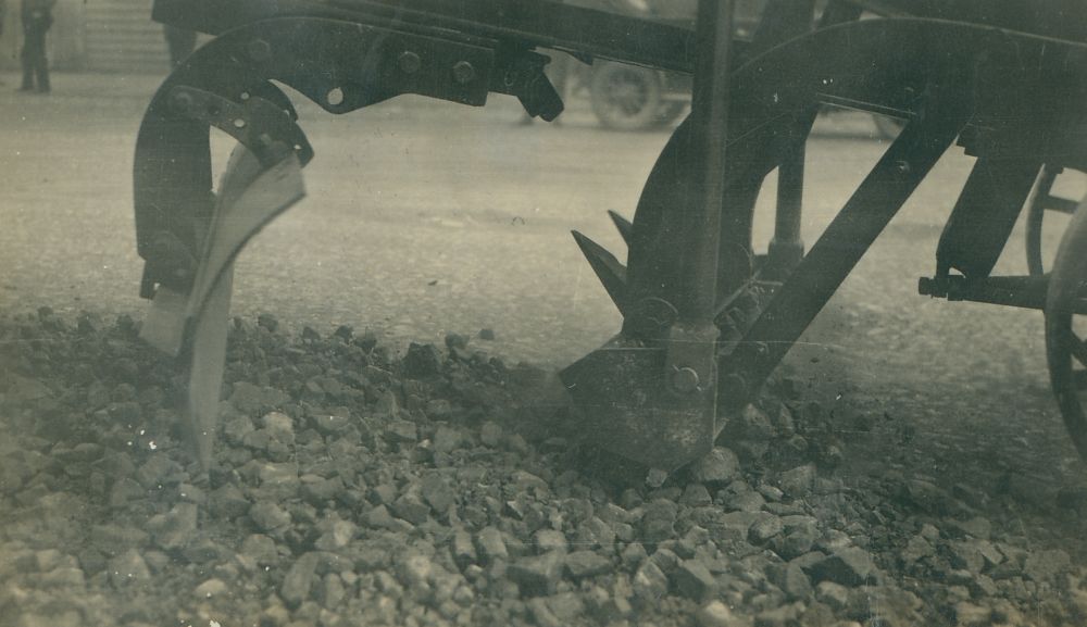 The underside of a grader tilling rocks along a street, ca. 1930