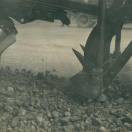 The underside of a grader tilling rocks along a street, ca. 1930